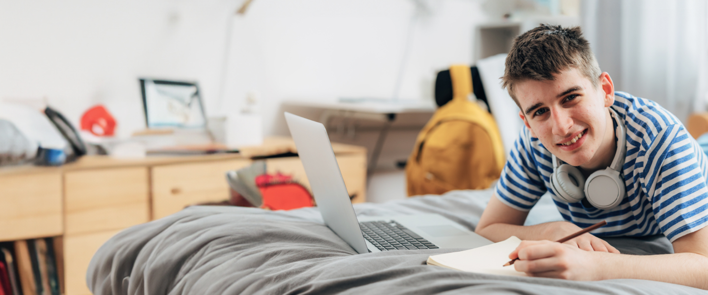 A smiling young person lies on a bed with grey sheets, wearing a blue and white striped shirt and white headphones around their neck. They are writing in a notebook and have an open laptop in front of them. The room has a desk, shelves, and a yellow backpack in the background.