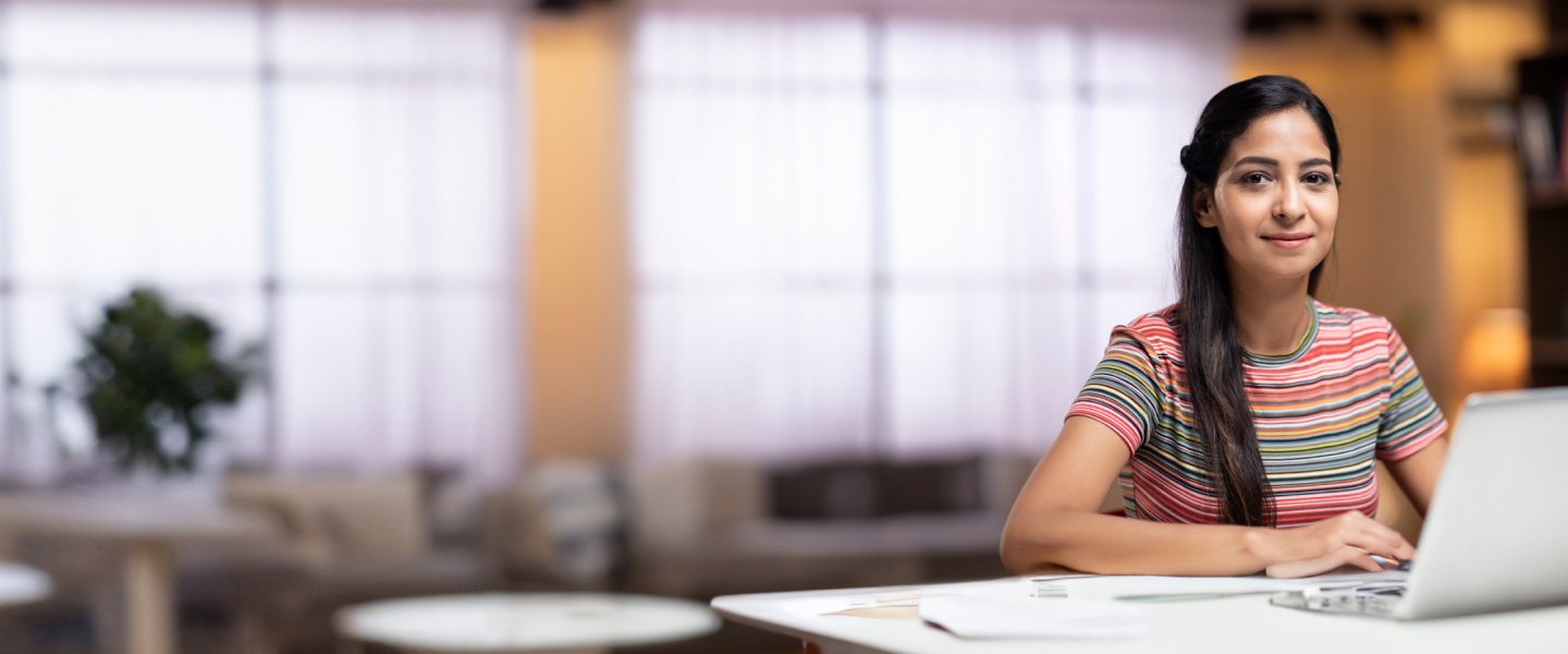 A woman wearing a striped shirt is sitting at a desk with a laptop in front of her. She is smiling and looking at the camera. The background features a softly lit modern interior with large windows and blurred furnishing.
